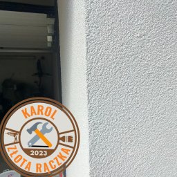 Złota Rączka Olsztyn -Karol - Świeżo nałożony, biały tynk strukturalny na ścianie zewnętrznej, widoczny fragment ościeżnicy drzwi garażowych i logo firmy remontowej 'Karol Złota Rączka 2023'.