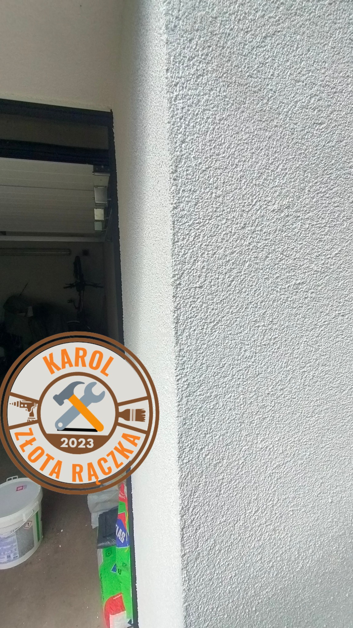 Świeżo nałożony, biały tynk strukturalny na ścianie zewnętrznej, widoczny fragment ościeżnicy drzwi garażowych i logo firmy remontowej 'Karol Złota Rączka 2023'.
