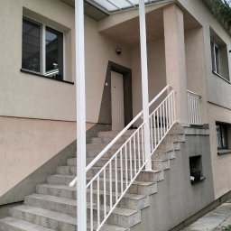 Arek Płoński - Zewnętrzne schody z jasnymi płytkami i białą metalową balustradą, prowadzące do wejścia domu z zadaszeniem. Widoczne okna i fragment elewacji w dwóch odcieniach.