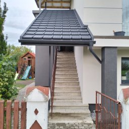 Arek Płoński - Zewnętrzne schody betonowe prowadzące do wejścia domu, z metalową balustradą i zadaszeniem z dachówki. W tle widoczny plac zabaw dla dzieci.