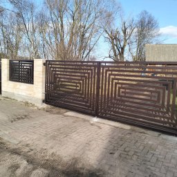 Arek Płoński - Nowoczesna, brązowa brama wjazdowa z geometrycznym wzorem, osadzona na jasnych, kamiennych słupkach. W tle drzewa bez liści i fragment budynku.