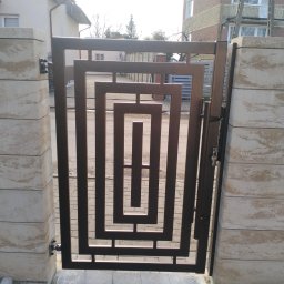 Arek Płoński - Nowoczesna, brązowa furtka metalowa z geometrycznym wzorem, zamontowana między dwoma kamiennymi słupkami. Widoczny fragment chodnika z kostki brukowej i budynki w tle.