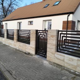 Arek Płoński - Nowoczesne ogrodzenie z metalowymi przęsłami o geometrycznym wzorze, osadzone na niskim murku z kamienia naturalnego. Brama w tym samym stylu. Dom w tle.