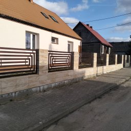 Arek Płoński - Nowoczesne ogrodzenie z metalowymi przęsłami o geometrycznym wzorze, zamontowane na niskim murku z płytek imitujących kamień, na tle domów i chodnika.