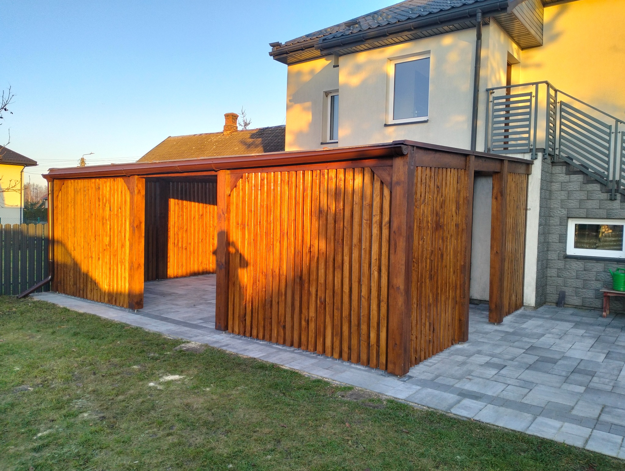 Drewniana pergola zadaszona, z pionowych desek, przylegająca do budynku z szarymi schodami zewnętrznymi. Kostka brukowa na podłożu, trawnik. Ciepłe światło słoneczne.