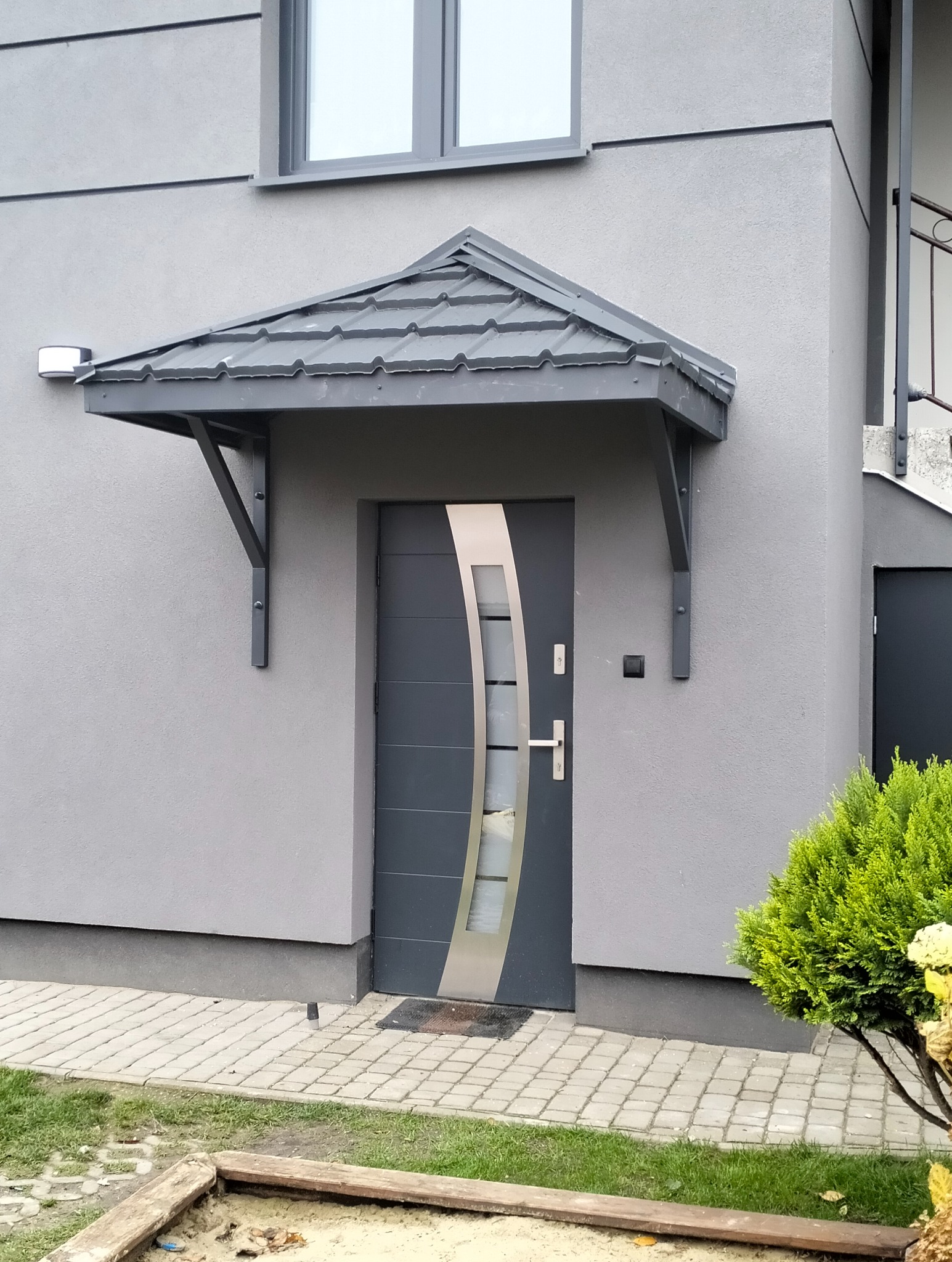 Szara elewacja domu z nowoczesnymi drzwiami wejściowymi i zadaszeniem z szarej dachówki, wspartym na metalowych wspornikach. Widoczny fragment chodnika i roślinności.
