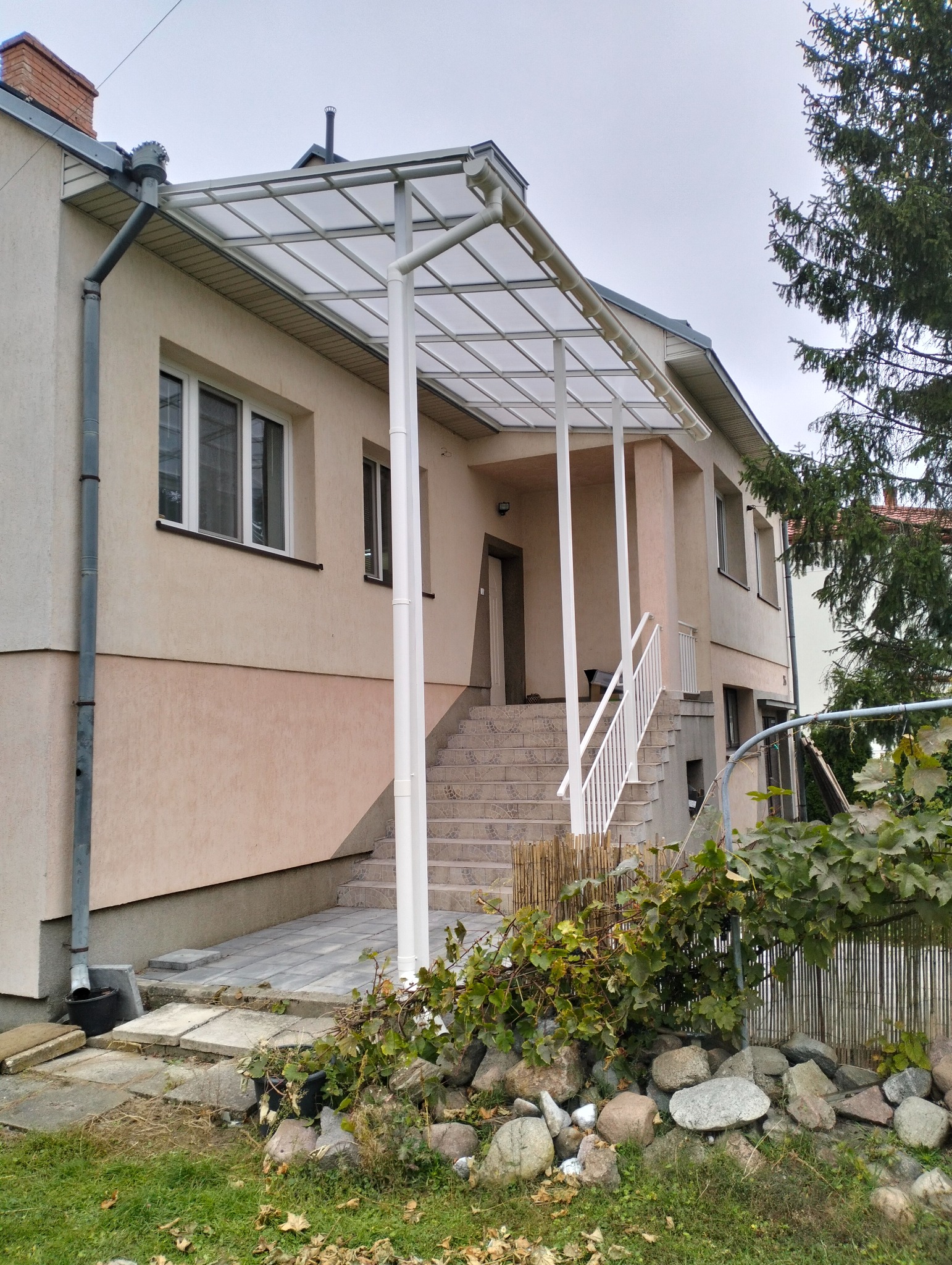 Zadaszenie nad schodami wejściowymi do domu, wsparte na białych kolumnach. Widoczne schody z jasnych płytek i biała balustrada. Tło stanowi elewacja budynku i fragment zieleni.