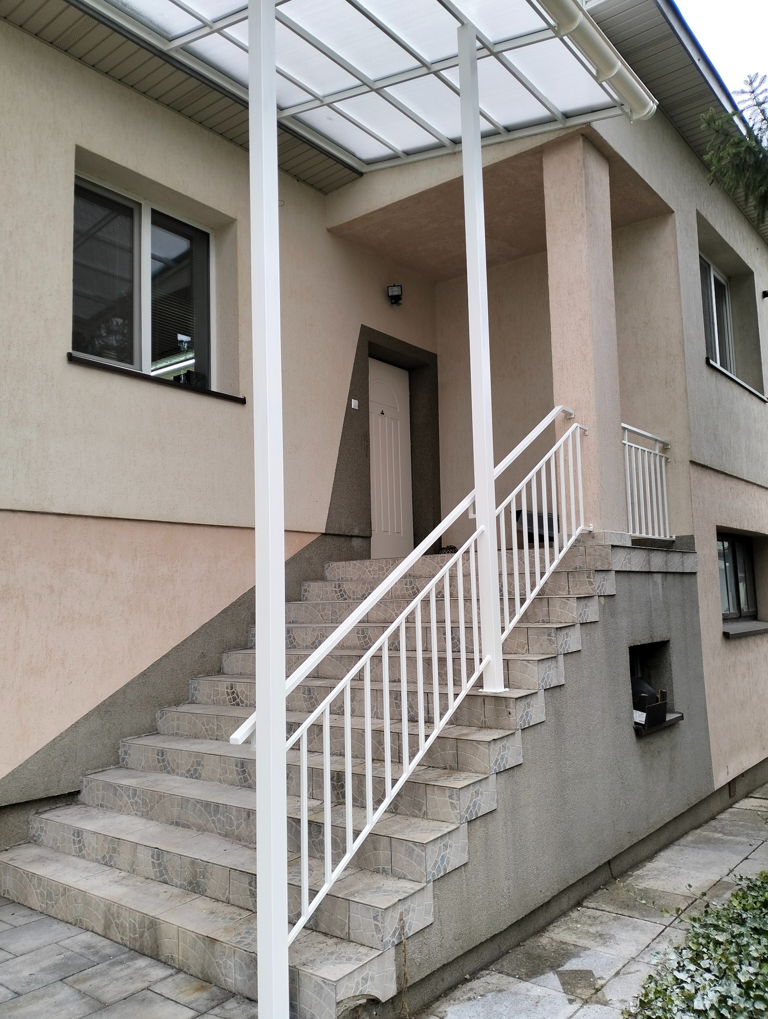Zewnętrzne schody z jasnymi płytkami i białą metalową balustradą, prowadzące do wejścia domu z zadaszeniem. Widoczne okna i fragment elewacji w dwóch odcieniach.