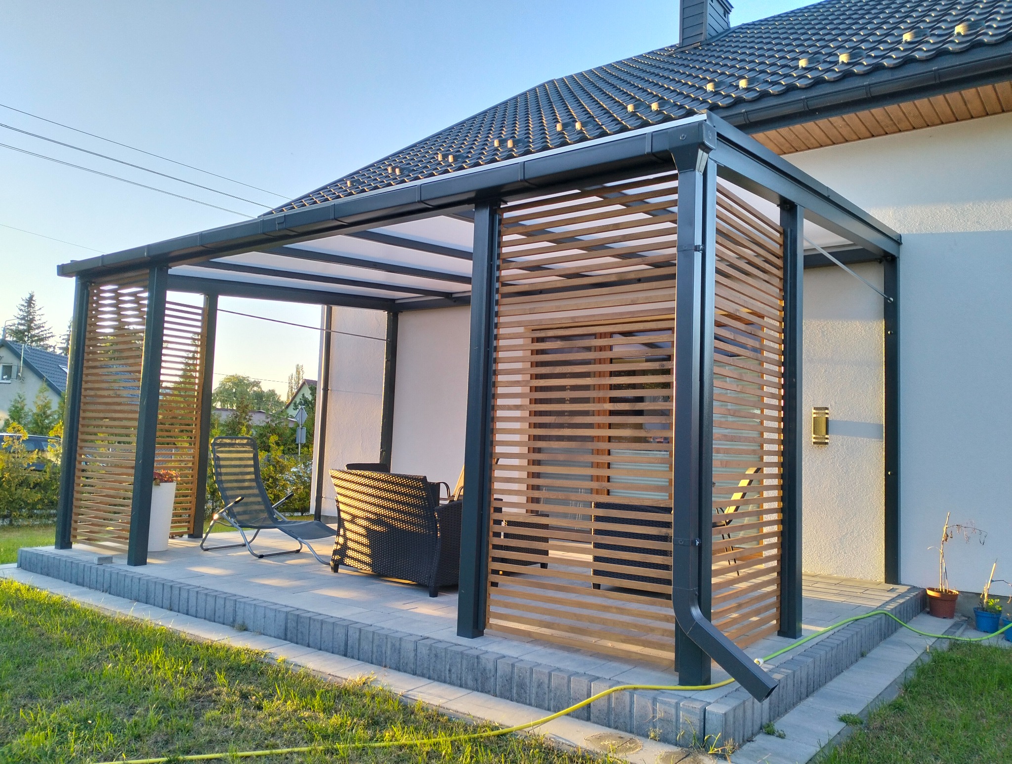 Nowoczesna pergola z metalową konstrukcją i drewnianymi lamelami, osłaniająca taras z meblami ogrodowymi. Widoczne detale wykończenia i oświetlenie.