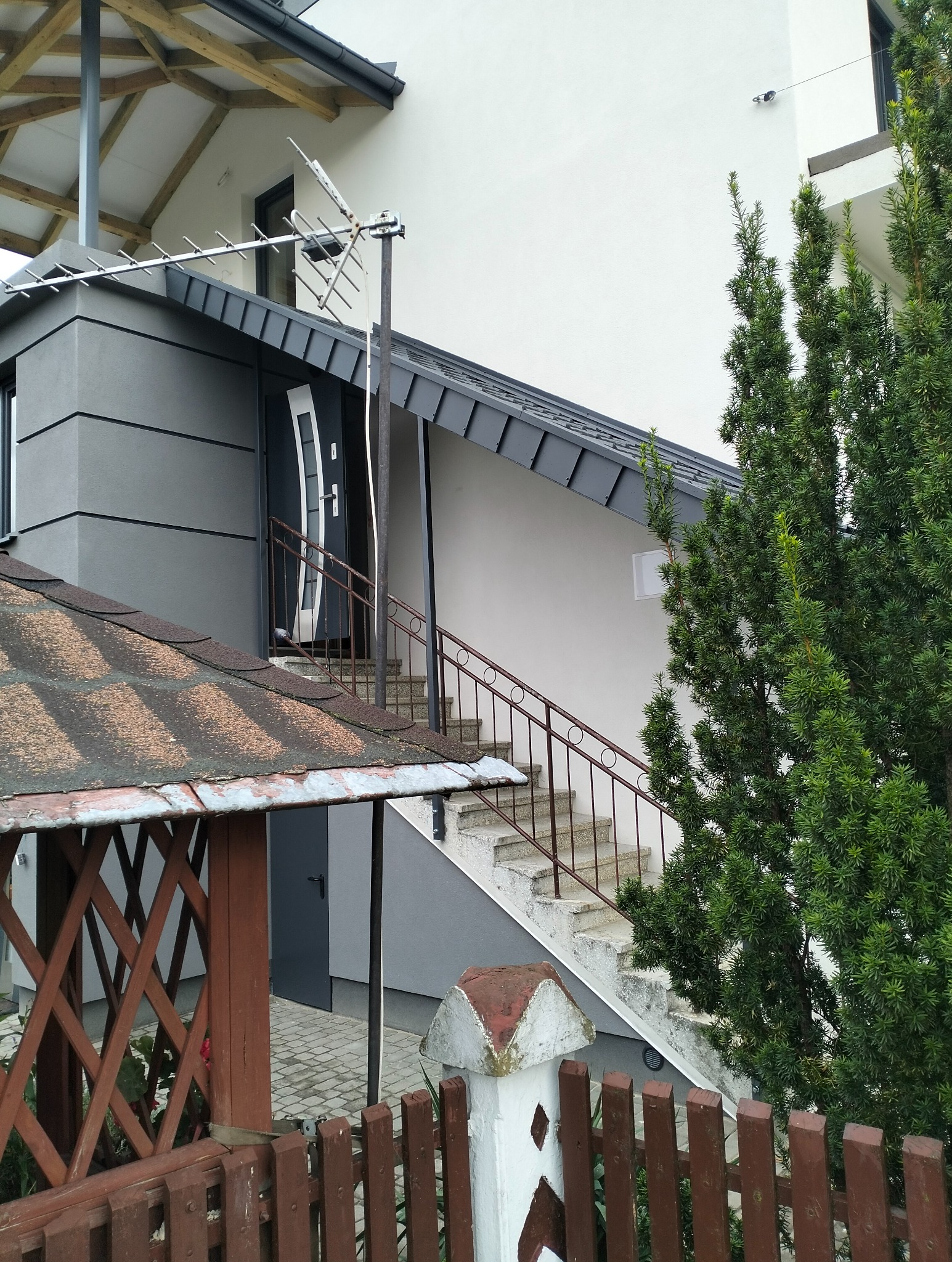 Zewnętrzne schody betonowe z metalową balustradą prowadzące do wejścia budynku, widoczne zza drewnianego ogrodzenia i fragmentu altany.