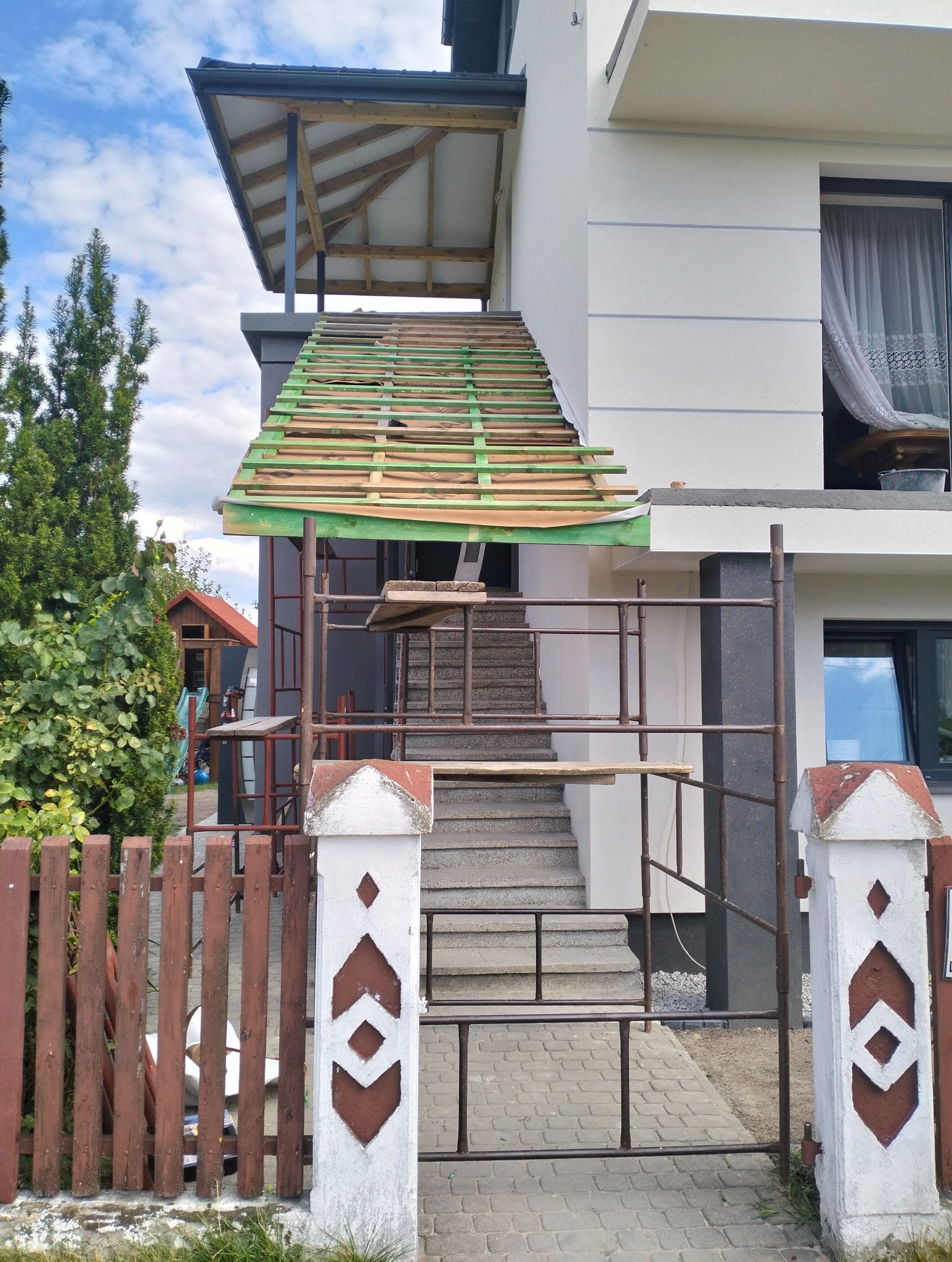Zadaszenie wejściowe w trakcie budowy, widoczne rusztowanie i drewniana konstrukcja dachu. Furtka z metalowymi prętami i słupki z dekoracyjnym wzorem.