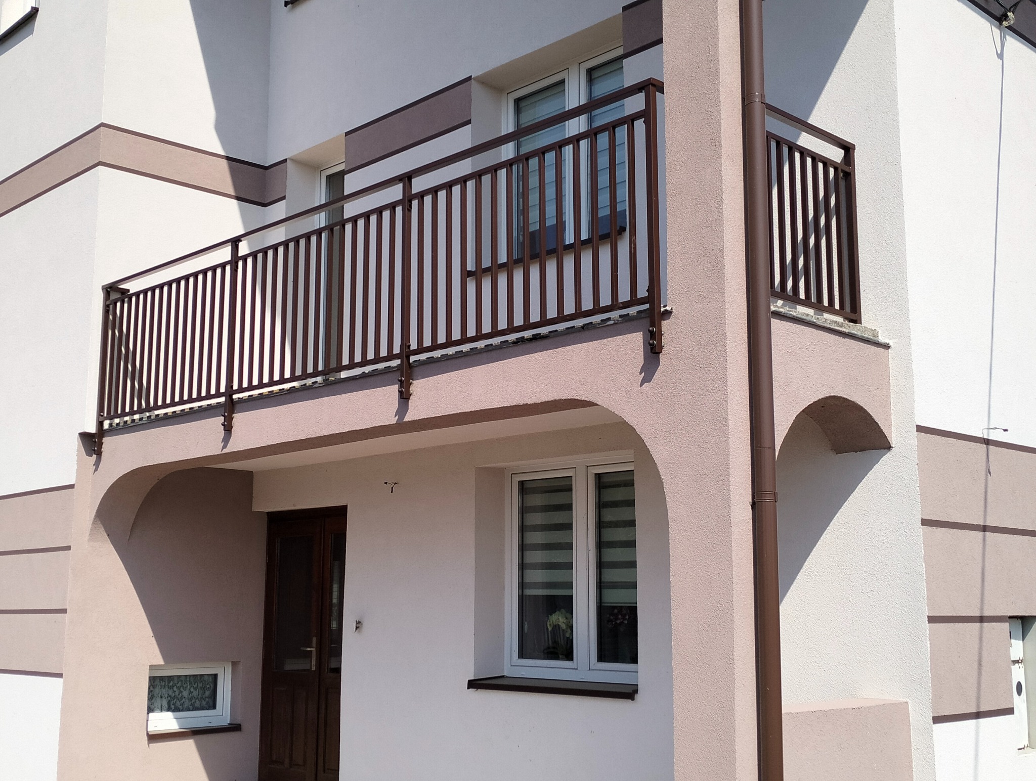 Balkon z brązową, metalową balustradą w nowoczesnym domu z jasną elewacją. Widoczne okna i drzwi wejściowe. Balustrada z pionowych elementów.
