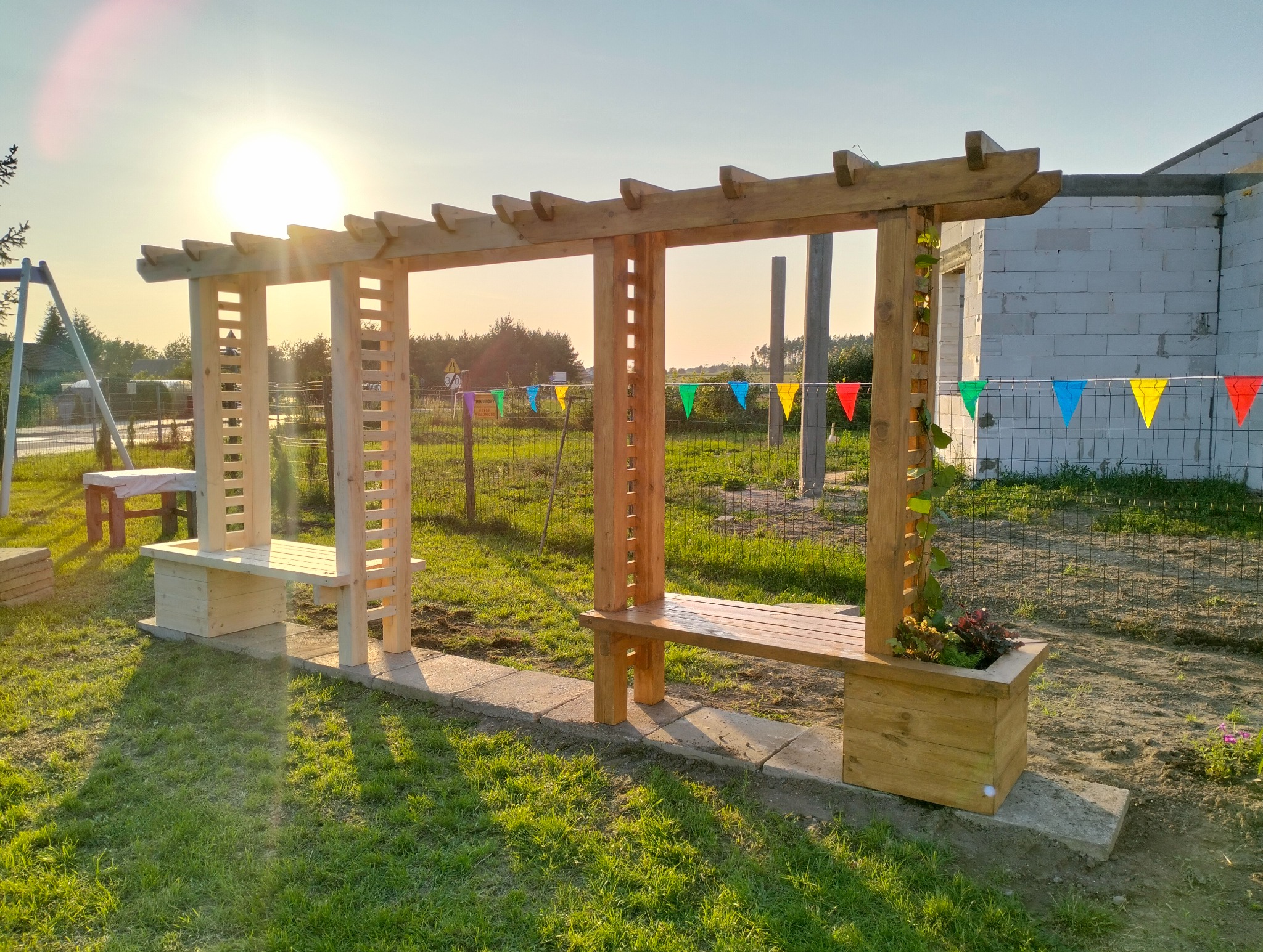 Drewniana pergola z ławkami i donicami, oświetlona promieniami zachodzącego słońca. W tle widoczny plac zabaw i budynek w trakcie budowy.