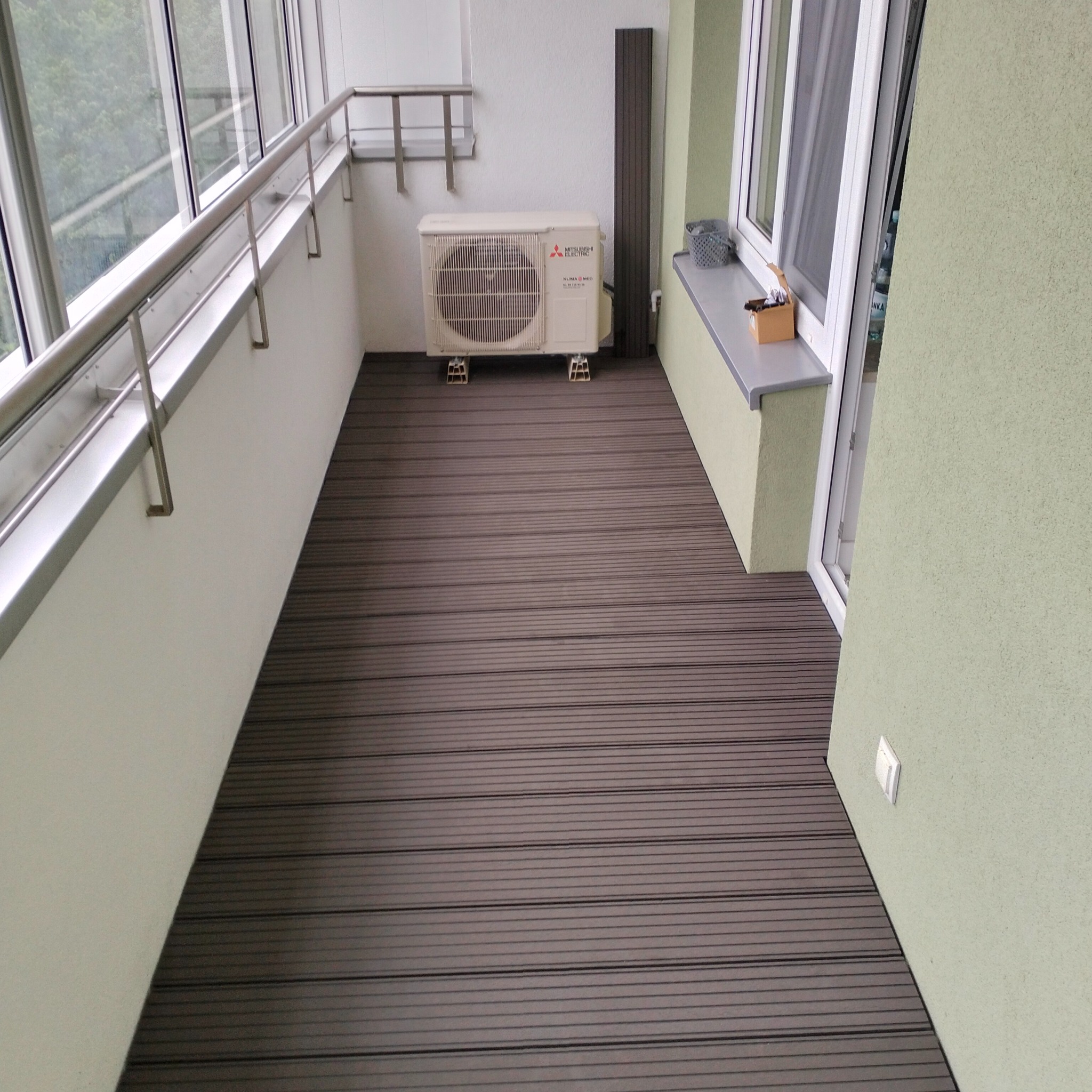 Balkon z brązową podłogą z desek kompozytowych, klimatyzatorem i metalową barierką. Widok na zewnątrz przez okna.