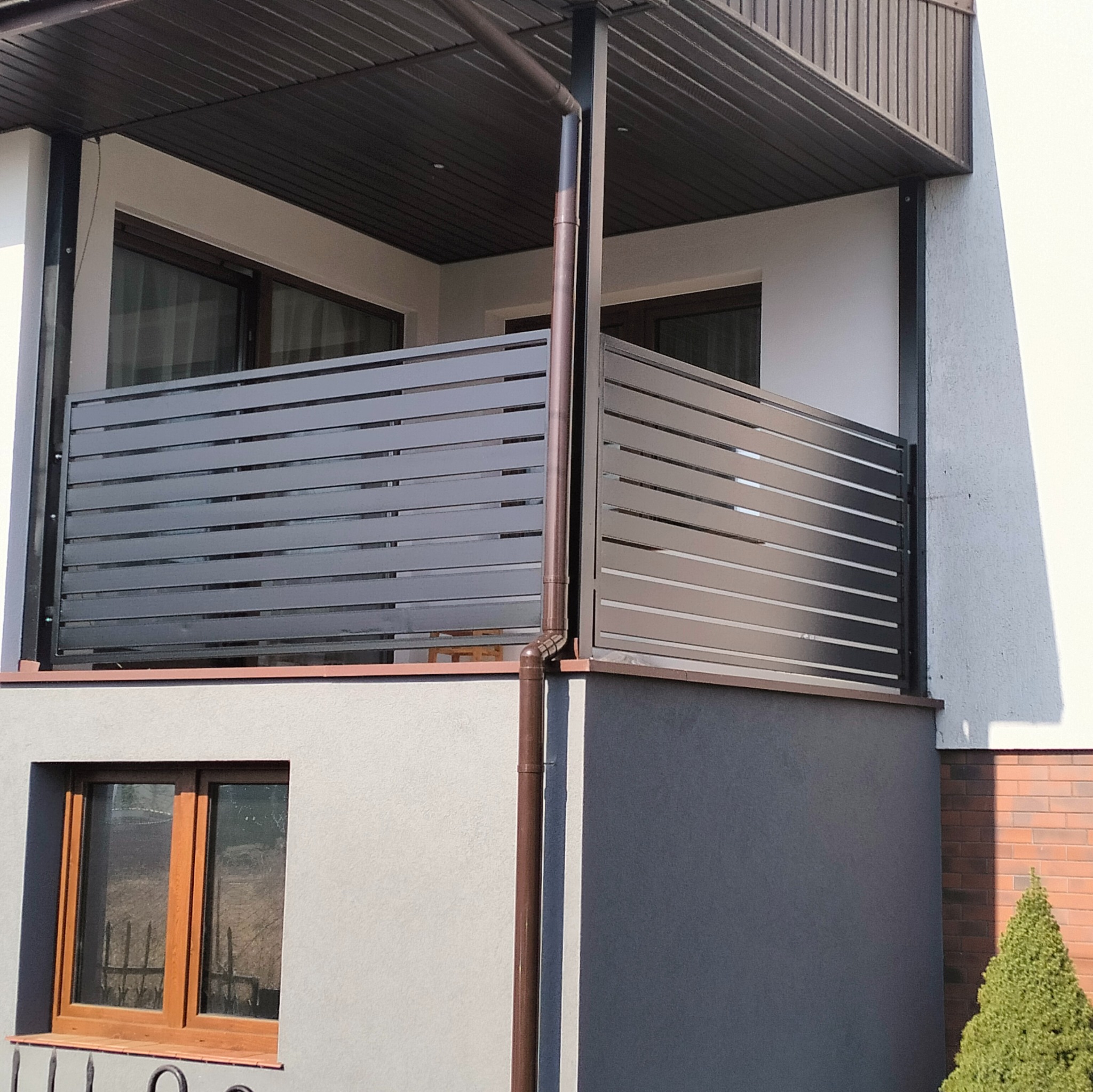 Nowoczesna zabudowa balkonu z poziomych, grafitowych paneli, widoczna z zewnątrz budynku. Eleganckie wykończenie, brązowa rynna, okno i fragment ceglanej ściany.