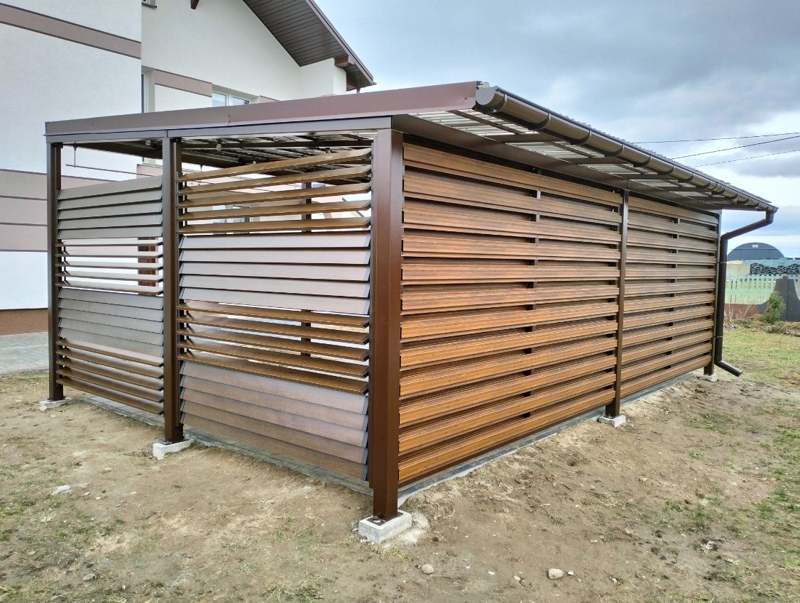 Nowoczesna pergola z brązowymi lamelami, betonowe podstawy słupów, widok z zewnątrz, na tle domu i pochmurnego nieba.