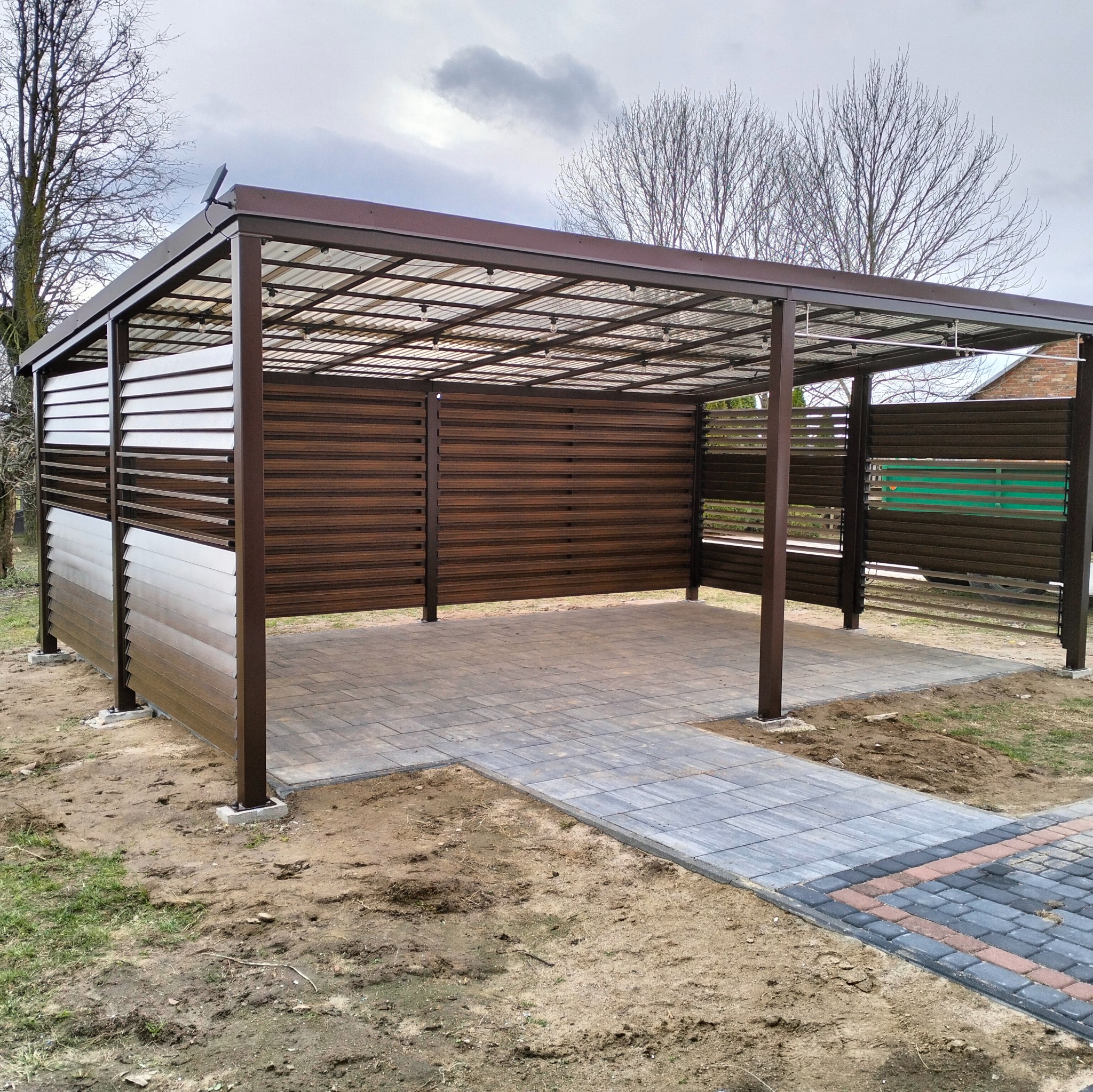 Nowoczesna pergola z brązowymi panelami i przezroczystym dachem, oświetlona lampkami, na tle szarej kostki brukowej i trawnika. Widok z zewnątrz, w pochmurny dzień.