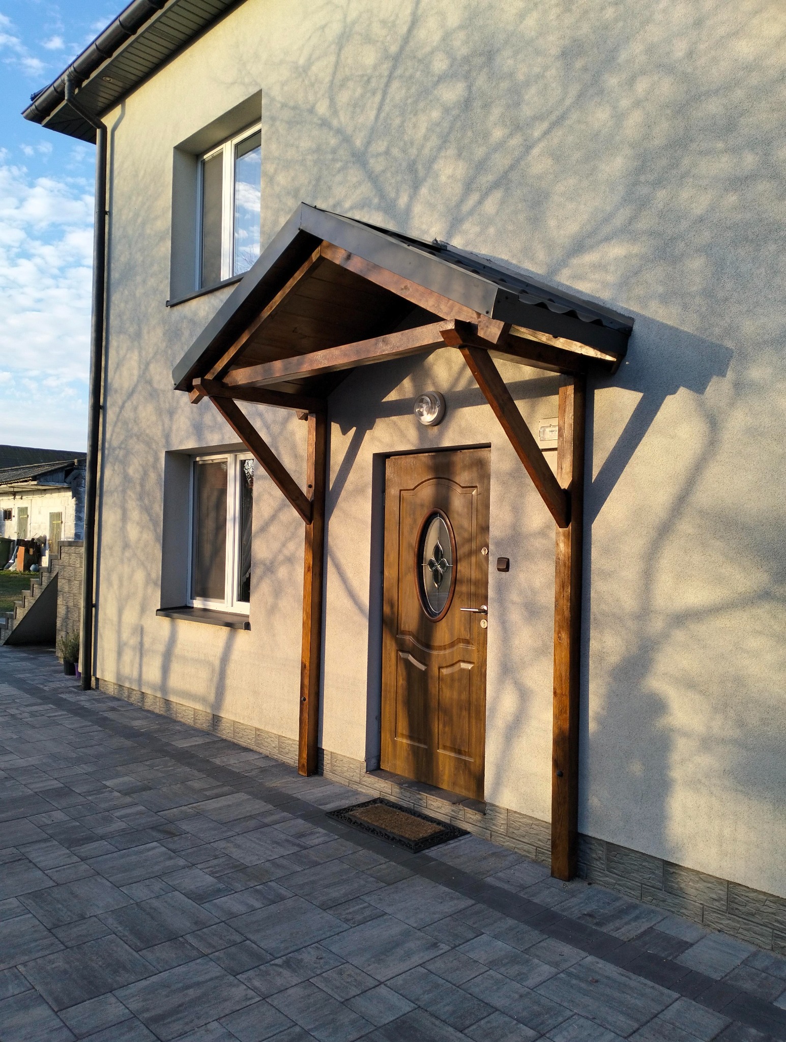 Drewniana, zadaszona weranda przy wejściu do domu, oświetlona popołudniowym słońcem. Widoczne okna i kostka brukowa. Cień drzewa na elewacji.