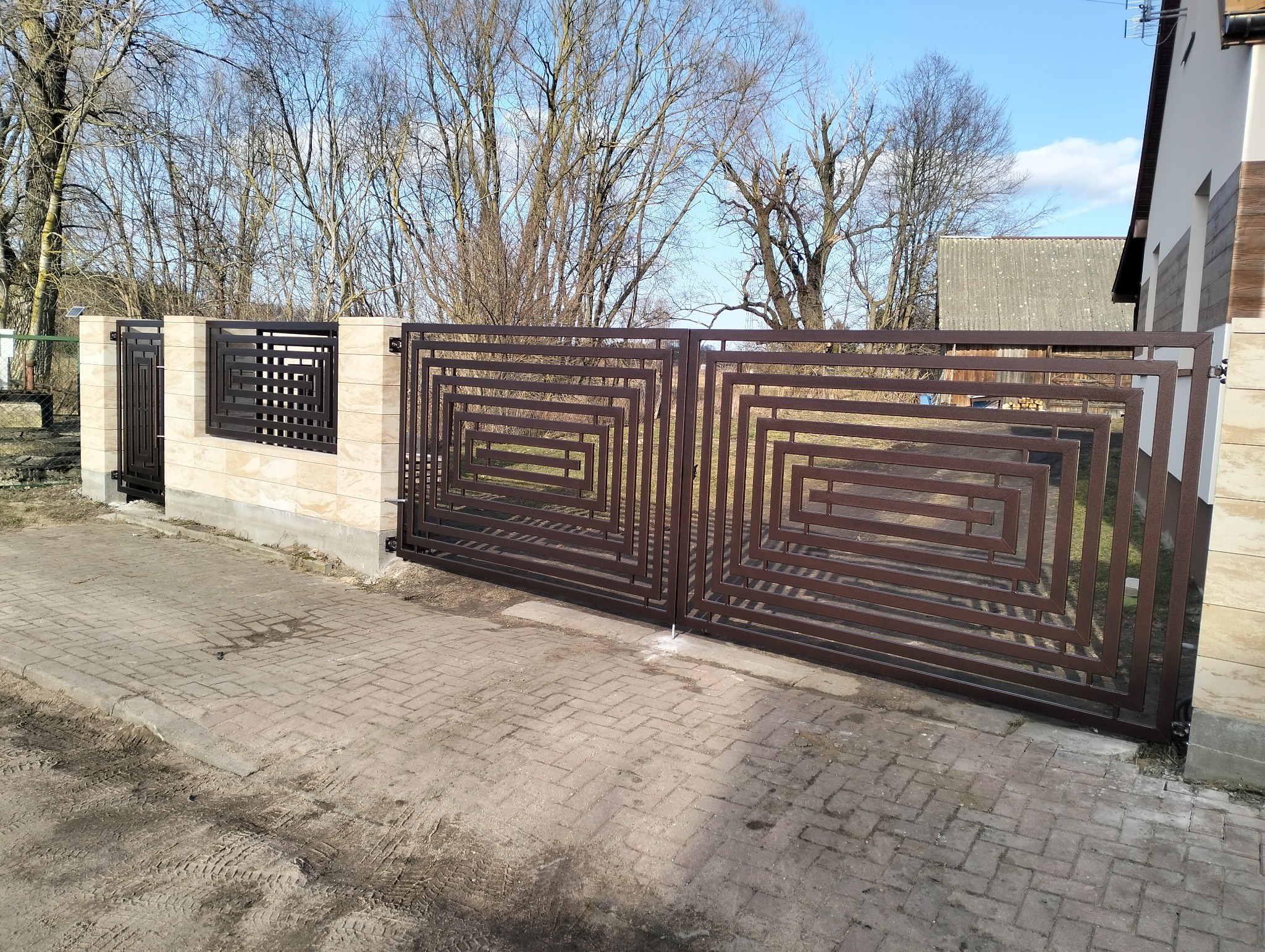 Nowoczesna, brązowa brama wjazdowa z geometrycznym wzorem, osadzona na jasnych, kamiennych słupkach. W tle drzewa bez liści i fragment budynku.