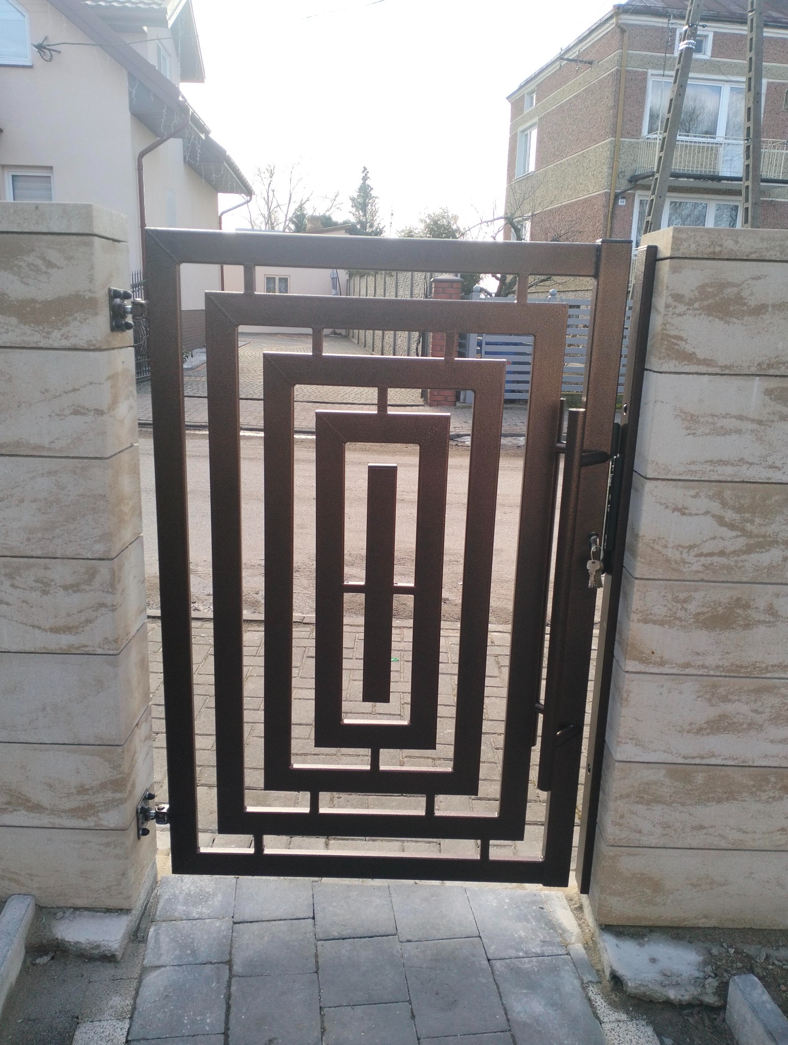 Nowoczesna, brązowa furtka metalowa z geometrycznym wzorem, zamontowana między dwoma kamiennymi słupkami. Widoczny fragment chodnika z kostki brukowej i budynki w tle.