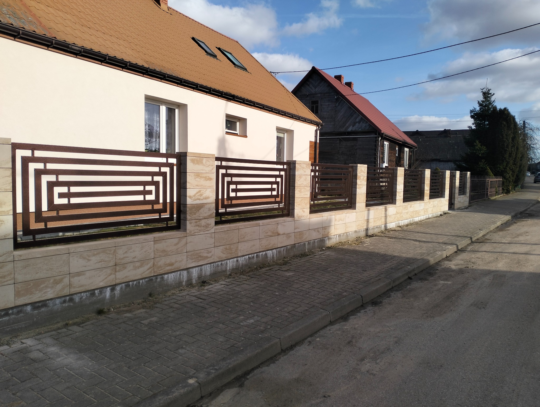 Nowoczesne ogrodzenie z metalowymi przęsłami o geometrycznym wzorze, zamontowane na niskim murku z płytek imitujących kamień, na tle domów i chodnika.