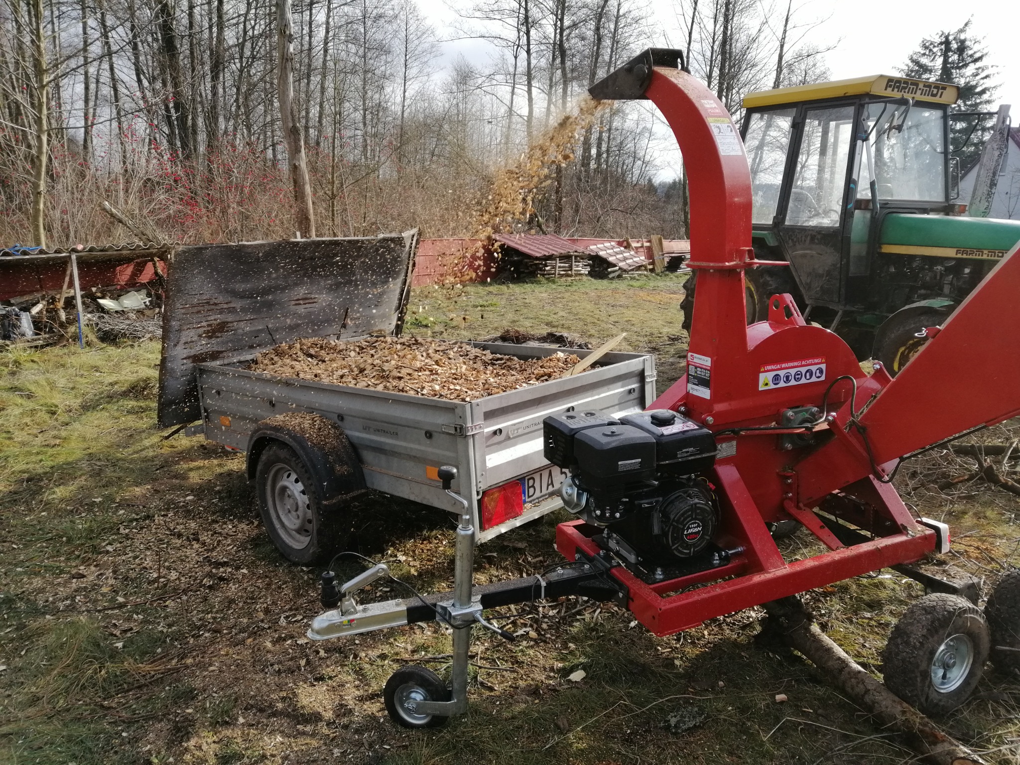 Czerwony rębak do gałęzi w trakcie pracy, wyrzucający zrębki do przyczepki, w tle traktor i drzewa bez liści.