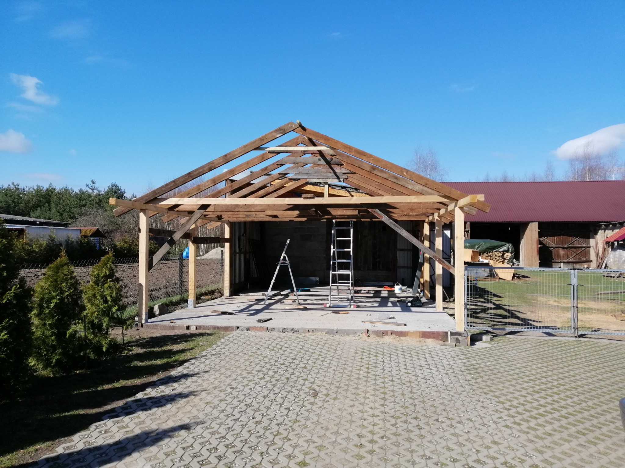 Konstrukcja drewniana z dachem w trakcie budowy, widoczne rusztowania i narzędzia, tło z zielenią i niebieskim niebem.