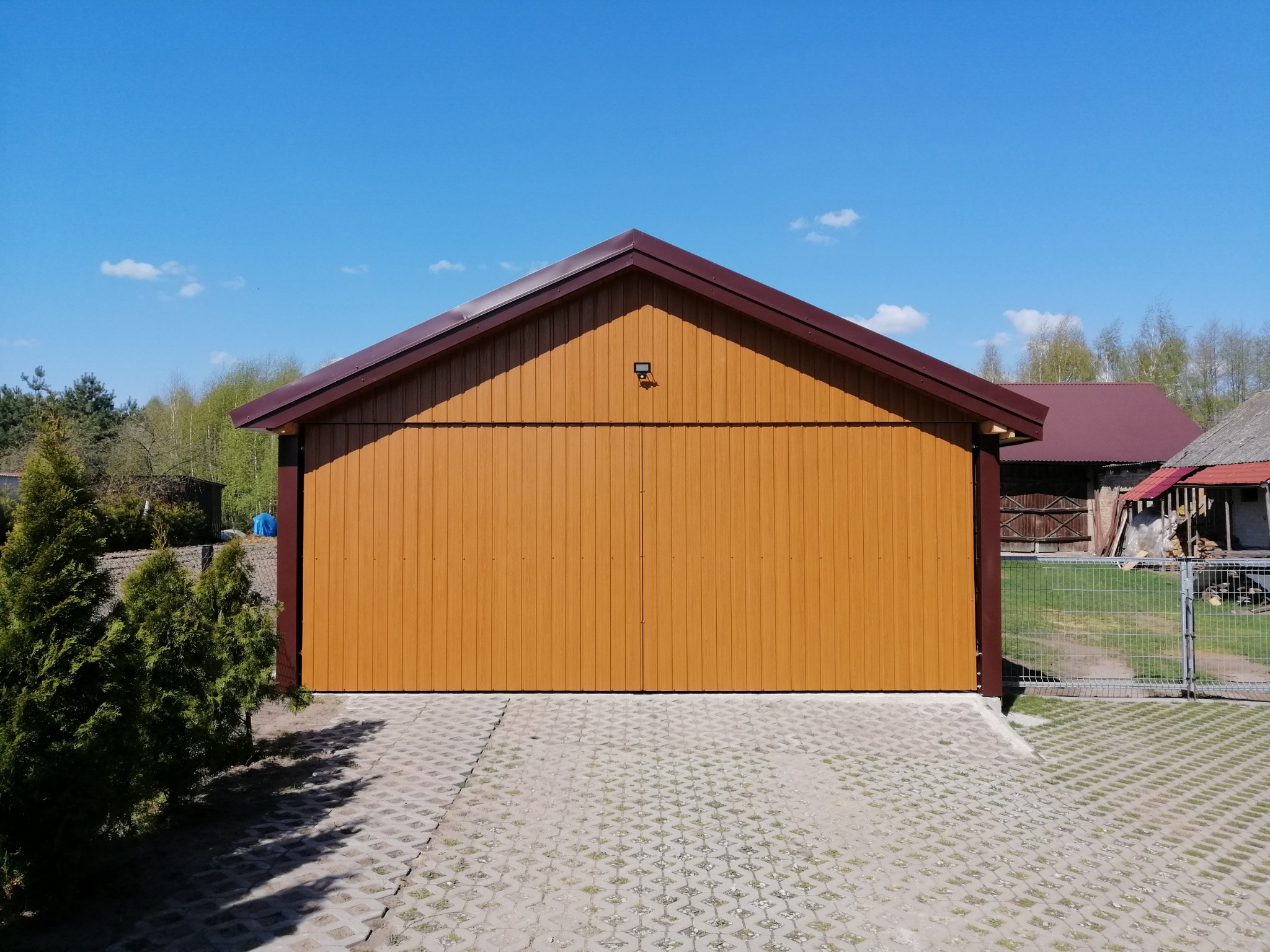 Nowoczesny, drewnopodobny garaż z brązowym dachem i elewacją, oświetlony światłem słonecznym, z podjazdem z ażurowych płyt betonowych, otoczony zielenią i błękitnym niebem.