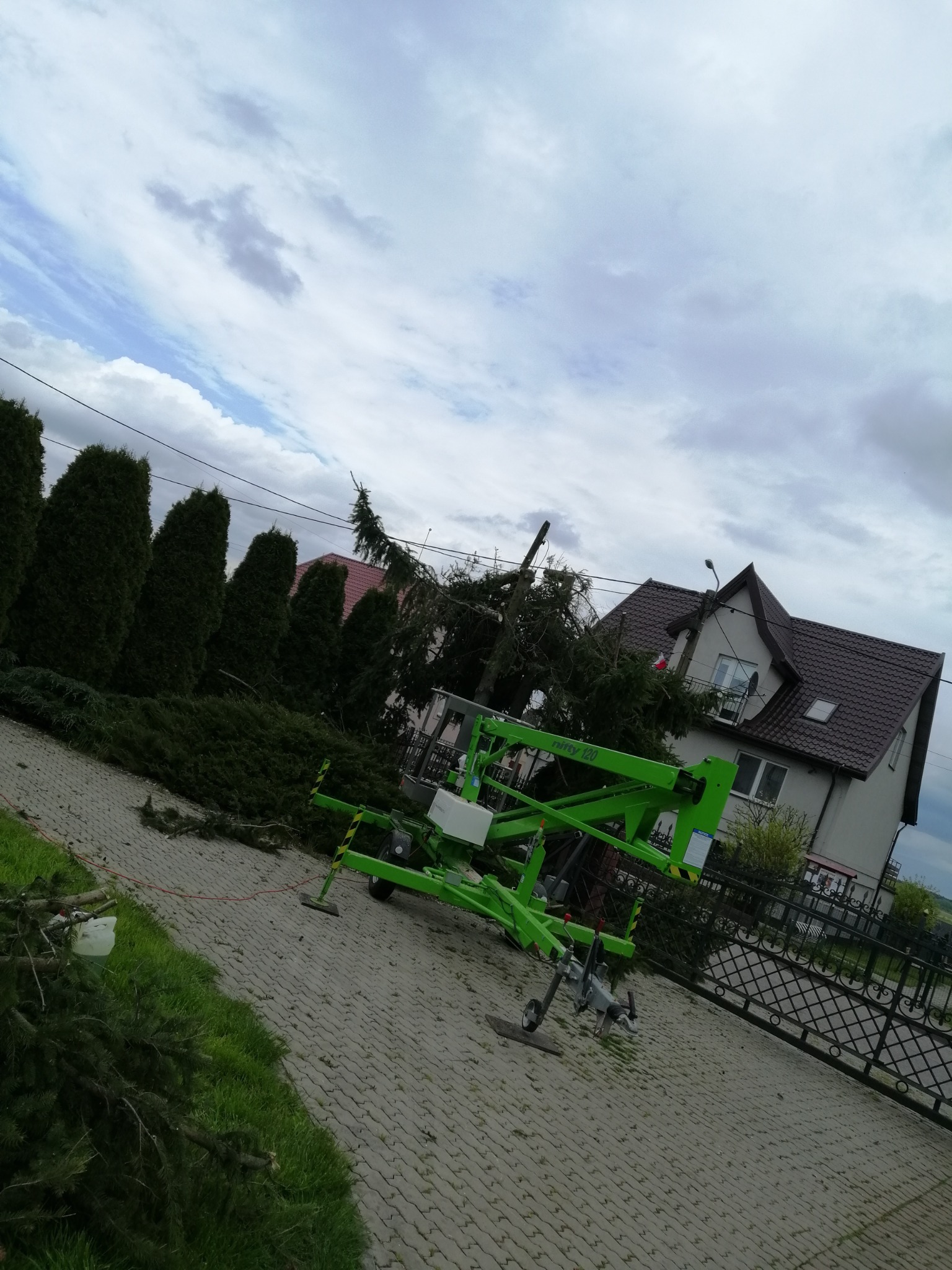Zielony podnośnik koszowy przy obciętej sośnie z porozrzucanymi gałęziami na kostce brukowej, w tle dom z brązowym dachem i wysokie tuje.