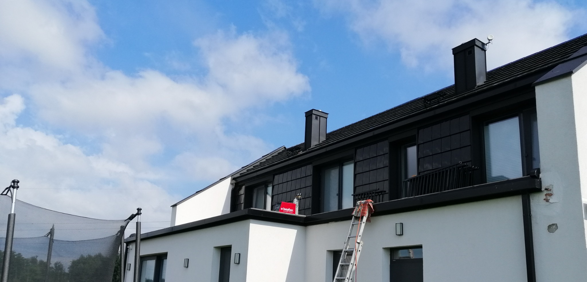 Dwupiętrowy dom z czarnym dachem i białymi ścianami, widoczna drabina oparta o dach, na dachu czerwony pojemnik z logotypem 'Bester', błękitne niebo z chmurami w tle.