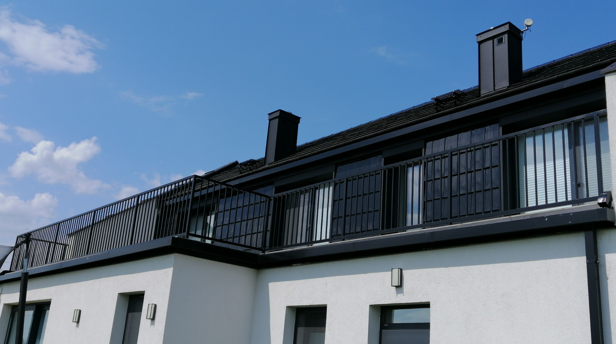 Nowoczesny budynek z białymi ścianami i czarnymi elementami wykończeniowymi, balkon z metalową balustradą na tle błękitnego nieba z delikatnymi obłokami.