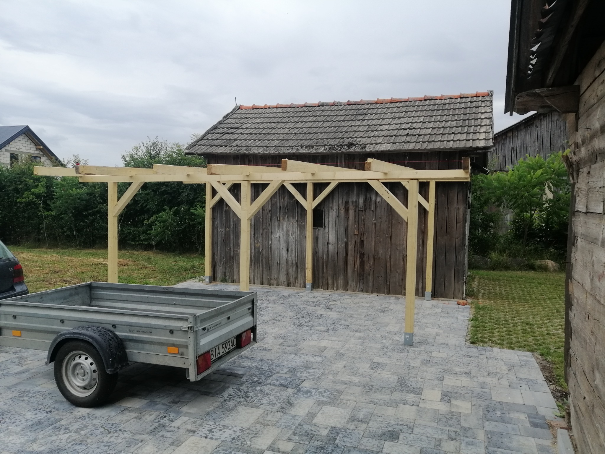 Surowa drewniana pergola w trakcie budowy, z przyczepką samochodową obok, na tle starego drewnianego budynku i kostki brukowej.