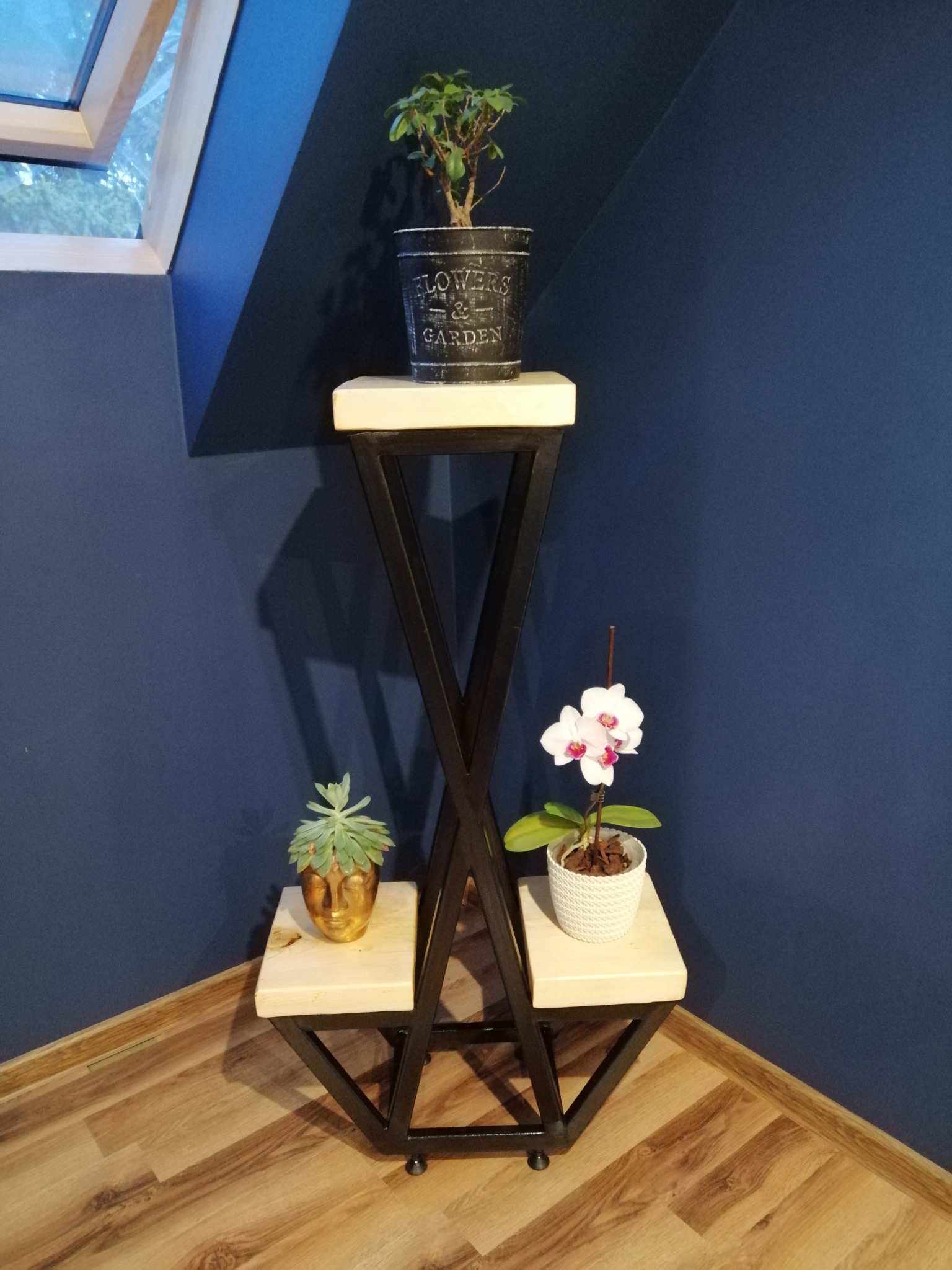 Trzypoziomowy kwietnik z metalu i drewna z sukulentem w złotej doniczce w kształcie głowy, bonsai w metalowej doniczce z napisem i białą orchideą w plecionej doniczce na tle granatowej ściany...