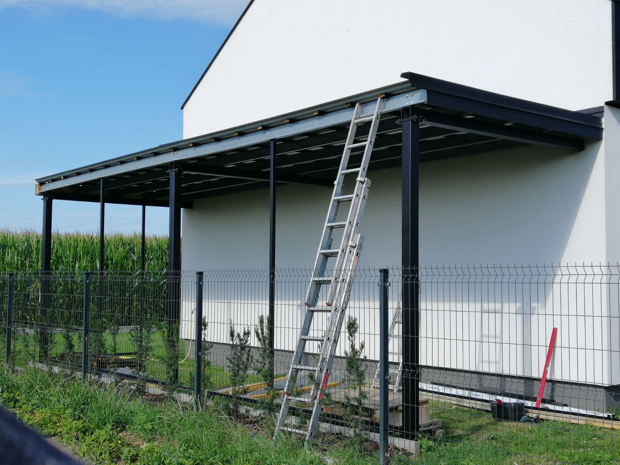Metalowa pergola w trakcie montażu przy białym domu, widoczna drabina oparta o konstrukcję, w tle pole kukurydzy i ogrodzenie panelowe.