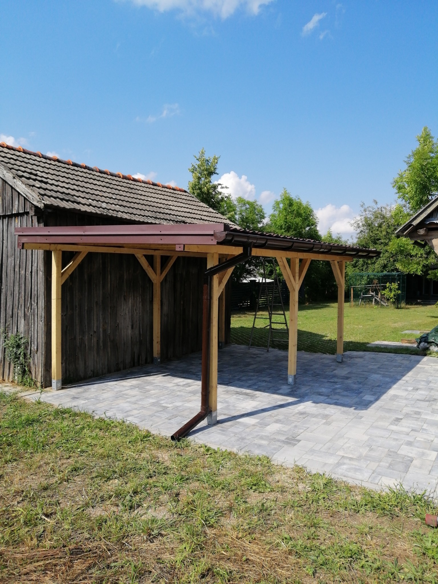 Zadaszenie z drewna nad szarym brukowanym tarasem, z rynną odprowadzającą wodę, przylegające do drewnianego budynku gospodarczego z czerwoną dachówką, w tle zielony trawnik i drzewa pod błękitnym...