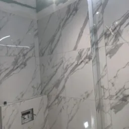 Ściana łazienki wyłożona białymi płytkami imitującymi marmur, z widocznym otworem pod instalację sanitarną i fragmentem sufitu z ukrytym oświetleniem LED.