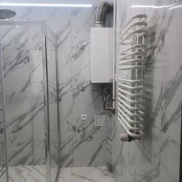 Nowoczesna łazienka z kabiną prysznicową, grzejnikiem drabinkowym i kotłem gazowym, wykończona płytkami imitującymi marmur z szarymi żyłami.