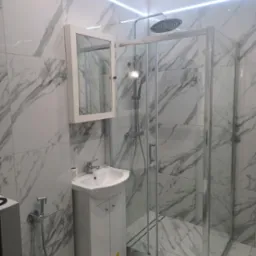 Nowoczesna łazienka wykończona marmurowymi płytkami, kabina prysznicowa ze szklanymi drzwiami, biała szafka pod umywalką i wisząca szafka z lustrem, oświetlenie LED na suficie.