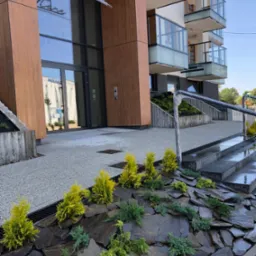 Nowoczesny ogród skalny przed budynkiem apartamentowym z żółtymi krzewami iglastymi i ciemnymi kamieniami łupkowymi, widoczne schody wejściowe i logo budynku.