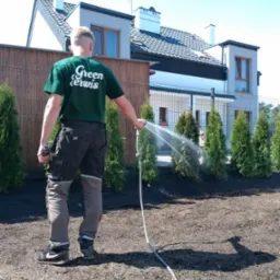 Mężczyzna w zielonej koszulce z logo firmy podlewa nowo posadzone tuje szmaragdowe wzdłuż ogrodzenia na tle nowoczesnego domu.