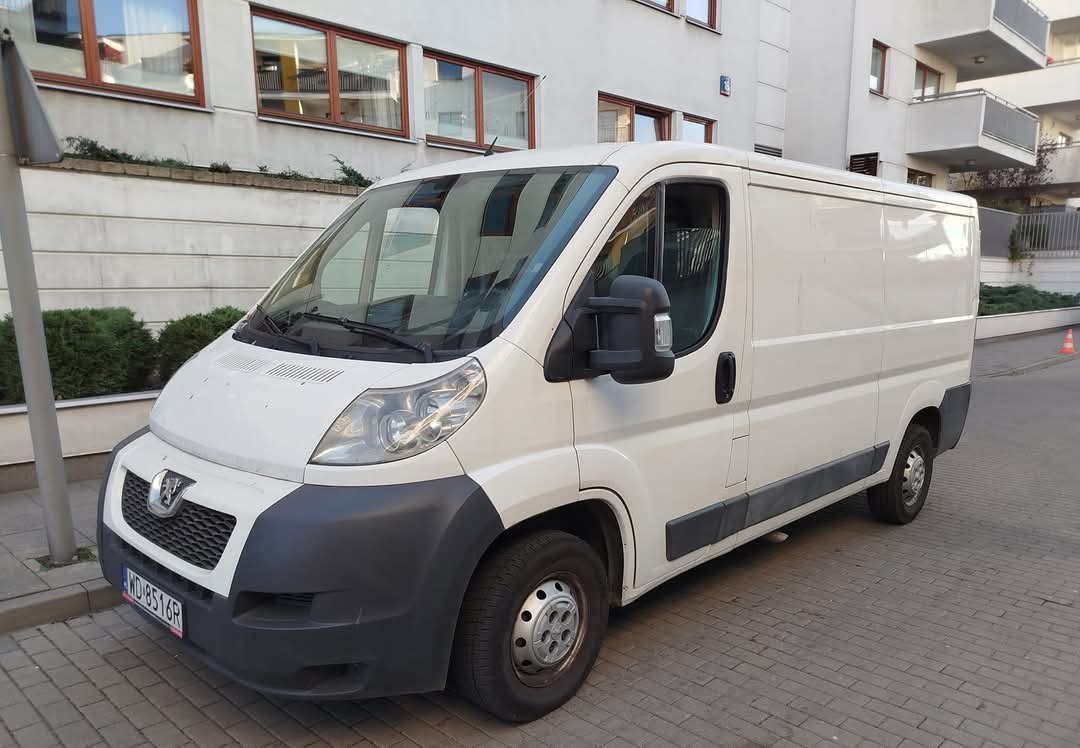 Biały Peugeot Boxer z numerem rejestracyjnym WD 8516R zaparkowany na chodniku z kostki brukowej na tle budynku mieszkalnego z brązowymi ramami okiennymi.