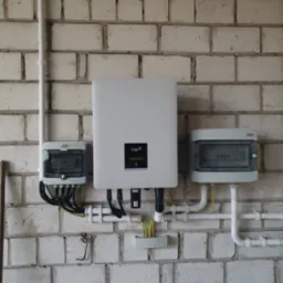 Instalacja elektryczna z białym inwerterem FoxESS na tle białej ściany z cegły, widoczne przewody, bezpieczniki i rury instalacyjne.