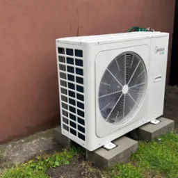 Biała zewnętrzna jednostka klimatyzacji Midea, umieszczona na betonowych blokach, obok brązowej ściany i czarnego śmietnika na zewnątrz budynku.