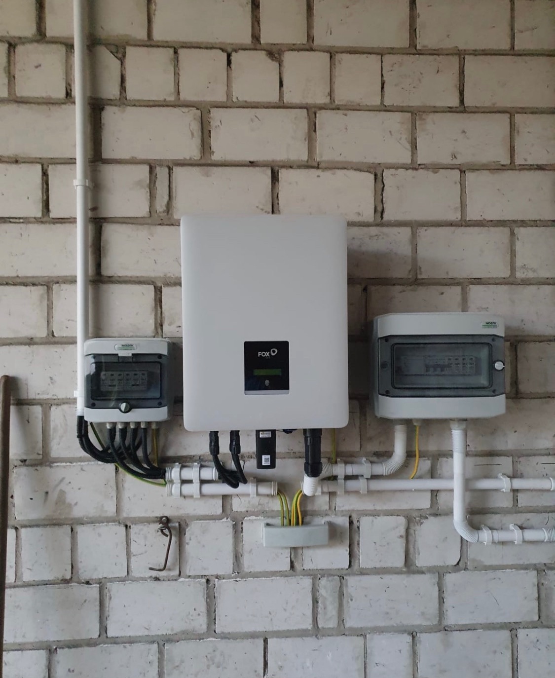 Instalacja elektryczna z białym inwerterem FoxESS na tle białej ściany z cegły, widoczne przewody, bezpieczniki i rury instalacyjne.