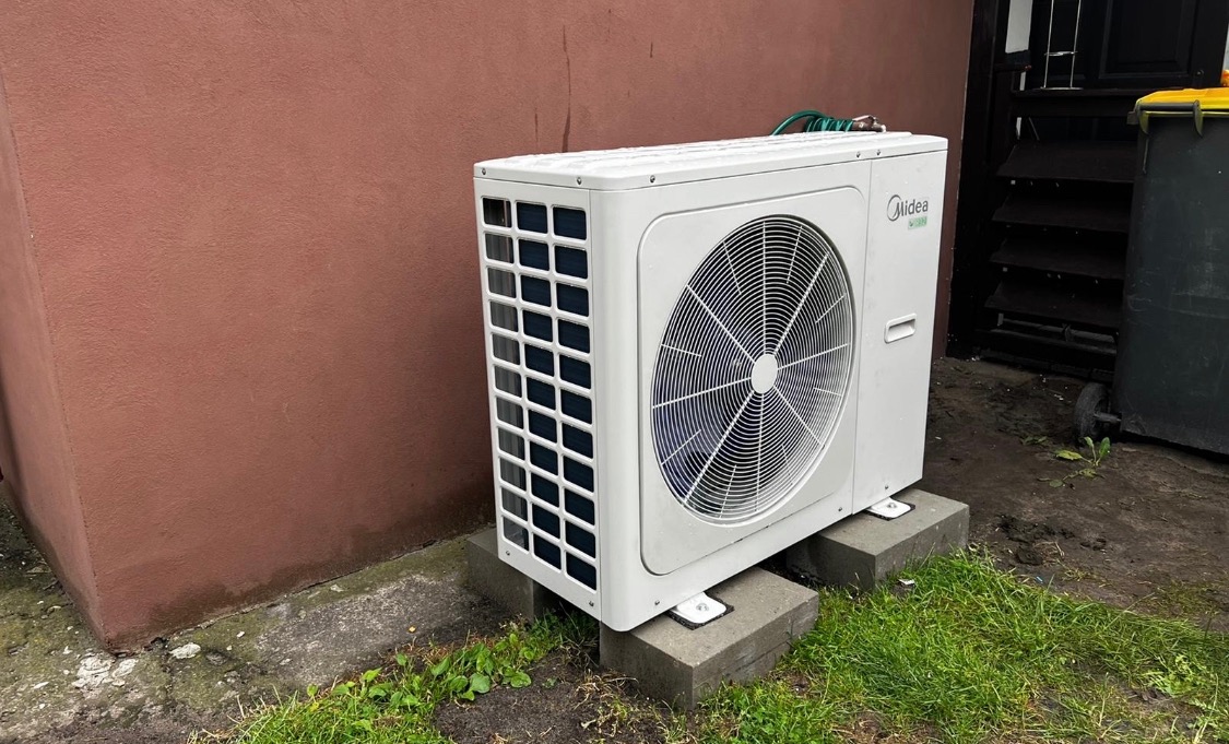 Biała zewnętrzna jednostka klimatyzacji Midea, umieszczona na betonowych blokach, obok brązowej ściany i czarnego śmietnika na zewnątrz budynku.