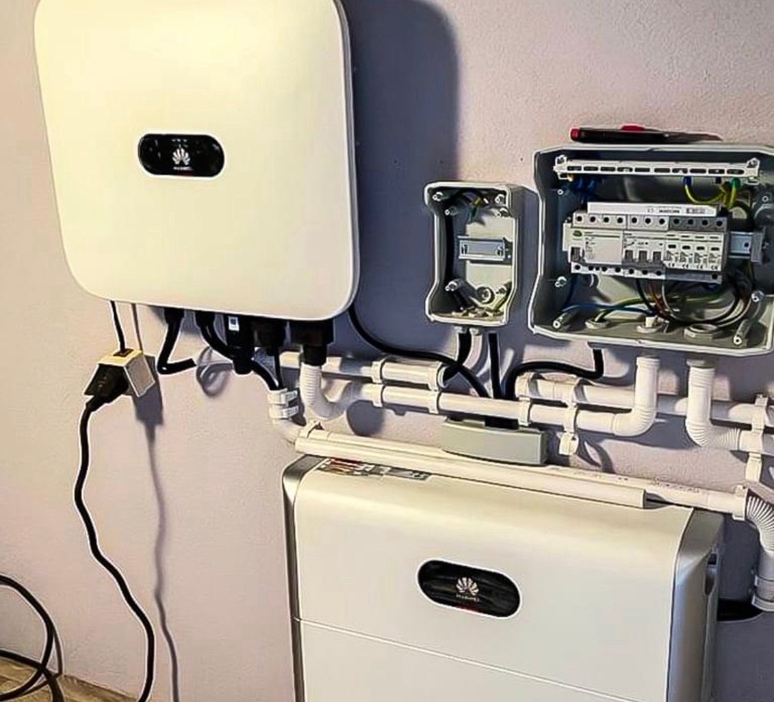 Biały inwerter fotowoltaiczny marki Huawei zamontowany na ścianie, z widoczną instalacją elektryczną i rurami PCV, obok otwartej skrzynki elektrycznej z bezpiecznikami i przewodami.