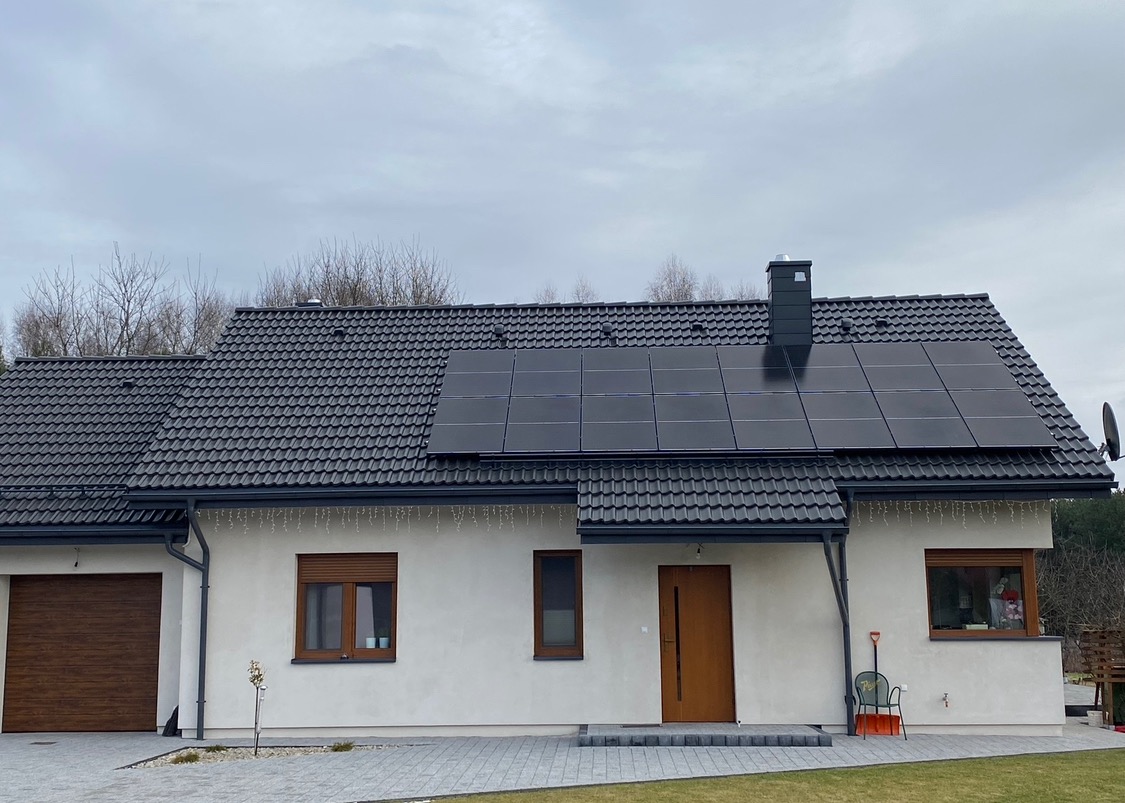 Dom jednorodzinny z zamontowanymi panelami fotowoltaicznymi na dachu z grafitowej dachówki, widoczne rynny, drewniane okna z roletami zewnętrznymi i kostka brukowa na podjeździe.