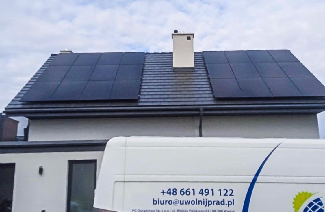 Panele słoneczne na dachu domu jednorodzinnego z szarej dachówki, widok z poziomu ulicy częściowo zasłonięty przez biały samochód dostawczy z numerem telefonu i adresem e-mail firmy.