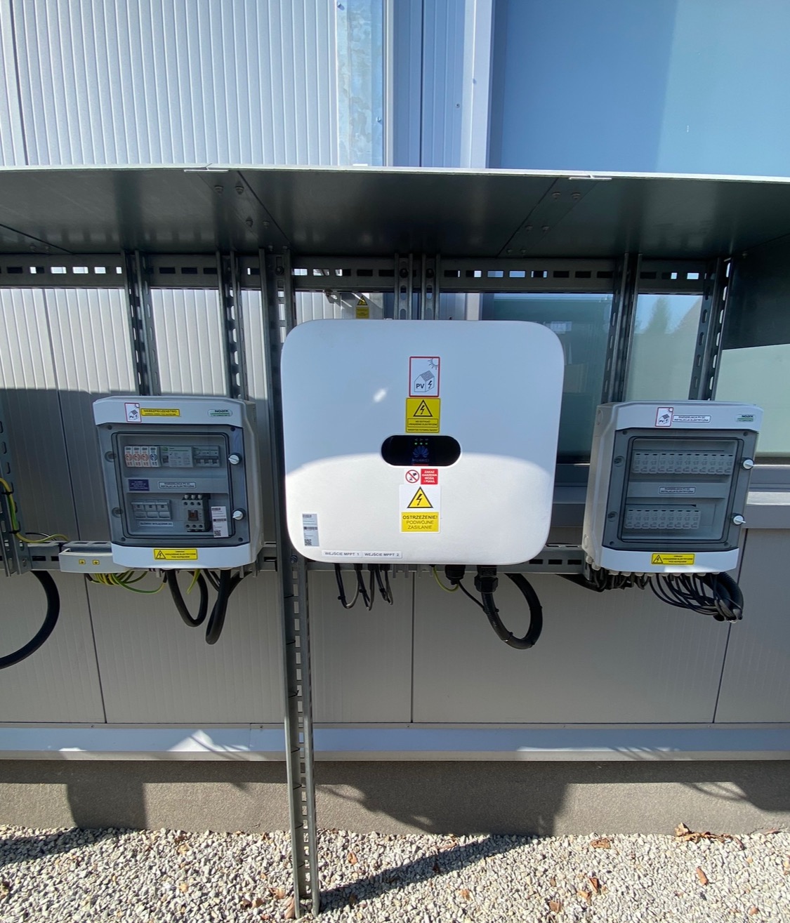 Biały falownik z widocznymi tabliczkami ostrzegawczymi i dwoma skrzynkami przyłączeniowymi zamontowany na szarej elewacji budynku z widocznymi przewodami.