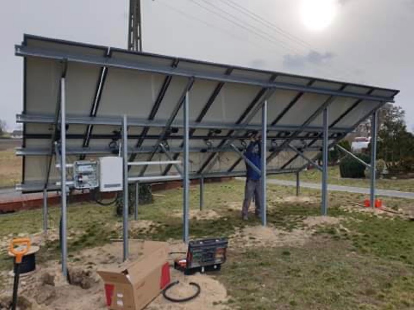 Montaż paneli słonecznych od strony konstrukcji wsporczej, widoczny pracownik w trakcie instalacji, skrzynka przyłączeniowa i narzędzia na ziemi.